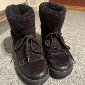 Svarta fluffiga boots - Säljer ett par svarta fluffiga boots i mycket bra skick. De har en mysig pälsliknande yta och snörning framtill. Perfekta för kalla vinterdagar när du vill hålla fötterna varma och bekväma. De har en robust sula som ger bra grepp. Storlek: 38. Nypris: 699 kr.