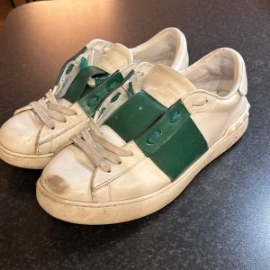 Valentino Garavani sneakers i vitt och grönt - Säljer ett par Valentino Garavani sneakers i vitt med gröna detaljer. Skorna har snörning och är i bra skick, men med några tecken på användning. Perfekta för en stilren och lyxig look. Kommer med originalförpackning. Varför jag säljer dom så billigt är att de är lite sönder längst fram?