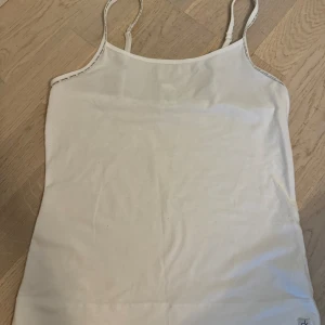Vit linnetopp från Calvin Klein - Säljer en stilren vit linnetopp från Calvin Klein. Den är ärmlös med smala axelband och har en diskret logga längs kanten. Perfekt för sommardagar eller som ett basplagg i garderoben. Materialet är mjukt och bekvämt, vilket gör den idealisk för både vardag och fest.