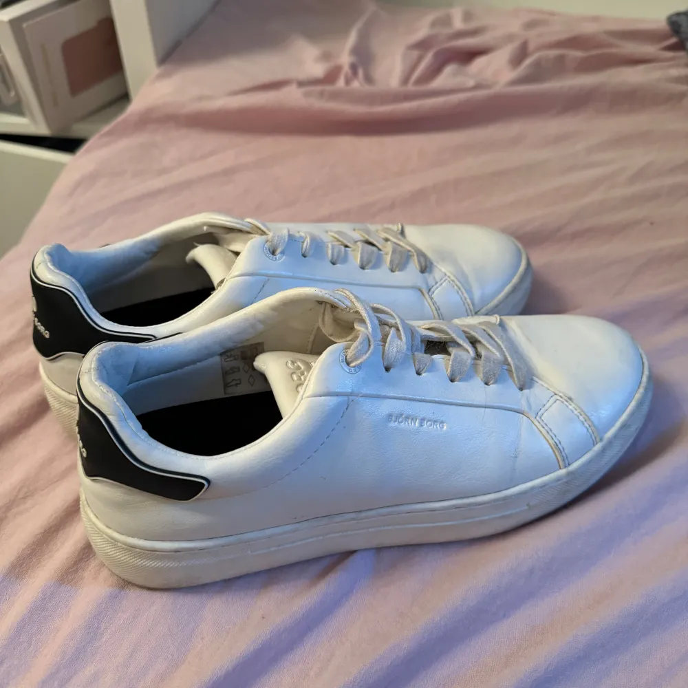 Säljer ett par vita sneakers från Björn Borg. De har en stilren design med svarta detaljer på hälen och snörning framtill. Skorna är i skinn och har en bekväm passform, perfekta för vardagsbruk.. Skor.