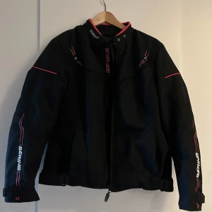 Svart motorcykeljacka från Bering - Säljer en svart motorcykeljacka från Bering, modell Lady Paradise. Jackan har en cool design med rosa och vita detaljer och Bering-loggan på ärmarna och ryggen. Den är långärmad och har en body fit-passform. Foder finns. Nyskick. 