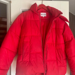 NAKD pufferjacka - En röd tjockare jacka från NAKD i storlek 36. Aningen större i storlek men de är en mild oversize variant. Avtagbar luva med dragkedja och två knappar för att knäppa ända upp till hakan. Dragkedja som försluter jackan och två fickor. Använd fåtal gån