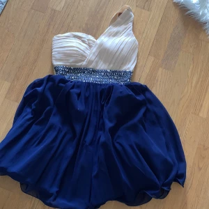Elegant klänning från Little Mistress - Säljer en superfin klänning från Little Mistress! Den har en one-shoulder design med en ljus överdel i plisserat material och en marinblå kjol. Midjan är dekorerad med ett glittrigt band som ger en extra touch av glamour. Perfekt för fest eller bal!