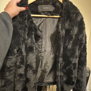 Svart pälsjacka från Vero Moda - Snygg och trendig svart pälsjacka från Vero Moda. Perfekt för att hålla sig varm och stilren under kyliga dagar. Jackan har en mjuk och fluffig yta som ger en lyxig känsla.