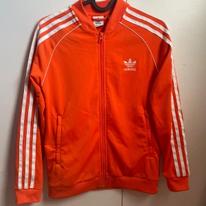 Orange träningsjacka från Adidas - Säljer en snygg orange träningsjacka från Adidas med de klassiska tre ränderna längs ärmarna. Jackan har dragkedja och två fickor fram. Perfekt för träning eller som en cool vardagslook. Passar både höst och vår!