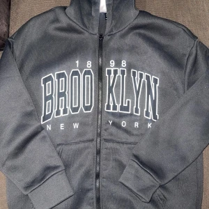 mörkgrå hoodie med brooklyn-tryck från Shein - säljer en mörkgrå hoodie med dragkedja och stort Brooklyn-tryck på framsidan från Shein. den har en mysig fleece-insida och en praktisk huva. erfekt för en avslappnad stil eller kyliga dagar. knappt använd!!