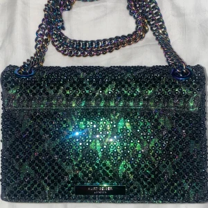 Glittrig axelväska med kedja - Snygg glittrig axelväska med regnbågsfärgad kedja och gnistrande stenar. Perfekt för en festkväll när du vill sticka ut. Väskan har en unik design med en dekorativ låsdetalj på framsidan.