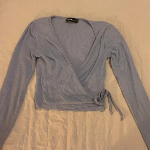 Ljusblå omlottopp från Fashion Nova - Säljer en superfin ljusblå omlottopp från Fashion Nova. Den är långärmad och har en snygg knytning i sidan. Perfekt för både vardag och fest! Toppen är i bra skick och passar perfekt till jeans eller kjol.