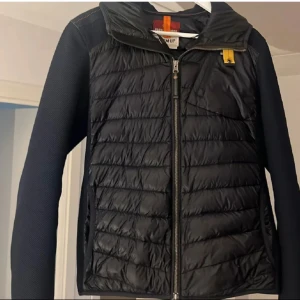 Svart jacka med huva och marinblå - Säljer en snygg svart jacka från Parajumpers. Den har en quiltad design med dragkedja och huva. Perfekt för höst och vår, med långa ärmar och en stilren look. Jackan är i mycket bra skick och passar både till vardags och lite finare tillfällen.