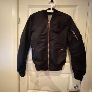 Svart bomberjacka från Alpha Industries - Säljer en snygg svart bomberjacka från Alpha Industries i storlek medium. Jackan har en klassisk design med rosa/guld dragkedja och fickor med knappar. Perfekt för höst och vår, och ger en cool look till vardagsoutfiten. Det ända som saknar på jackan är den röda grejen som brukar vara på ärmen