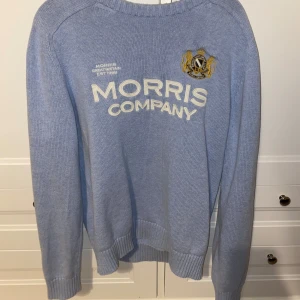 Ljusblå tröja från Morris - Säljer en snygg ljusblå tröja från Morris med broderad logga och text 'Morris Company' på framsidan. Tröjan är i mycket bra skick och har långa ärmar. Perfekt för både vardag och lite finare tillfällen. Passar bra till jeans eller chinos!