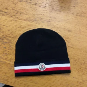Svart mössa  Moncler - Säljer en snygg svart mössa från Moncler i ull och kaninhår. Den har en cool randig detalj i rött, vitt och blått samt Moncler-loggan framtill. Perfekt för att hålla sig varm och stilren under kalla dagar. Tillverkad i Italien. Mössan är helt ny den är inte använt. Pris kan diskuteras
