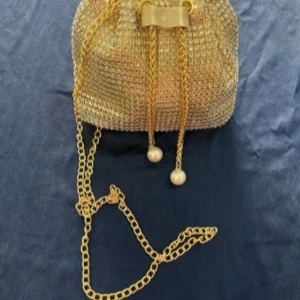 Guldig glittrig axelväska med pärldetaljer - Säljer en superfin guldig axelväska som verkligen glittrar! Den har en kedja som axelrem och söta pärldetaljer vid stängningen. Perfekt för fest eller nyår! Väskan är i syntetmaterial och har en elegant rosett på framsidan. Den är i mycket bra skick och redo att användas för att lysa upp vilken outfit som helst.