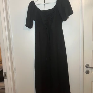 Svart långklänning med korta ärmar - Söt svart långklänning med korta ärmar och en smickrande offshoulder-design. Klänningen har en snygg snörning framtill som ger en fin detalj och en feminin touch.