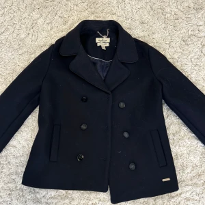Svart kappa från Massimo Dutti - Elegant svart kappa från Massimo Dutti i ullblandning. Klassisk design med dubbelknäppning och stilrena detaljer. Tyvärr är ena knappen på ärmen borta. Fint skick andvänd få tal gånger. 