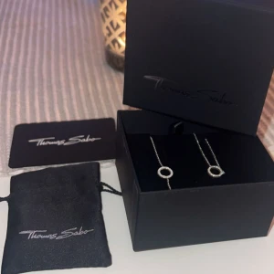 Silverhalsband från Thomas Sabo - Elegant silverhalsband med tillhörande armband från Thomas Sabo med en cirkulär hängsmycke dekorerad med glittrande stenar. Perfekt för att ge en touch av elegans till vilken outfit som helst. Levereras i originalförpackning. Aldrig använda🥰paketpris 1000kr för båda! 