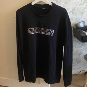 Svart långärmad tröja från J.Lindeberg - Snygg svart långärmad tröja från J.Lindeberg i storlek XL med tryck på framsidan. Perfekt för höst och vinter. Tillverkad i Portugal.