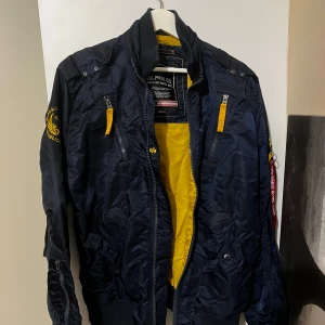 Mörkblå bomberjacka från Alpha Industries - Säljer en snygg mörkblå bomberjacka från Alpha Industries i storlek medium. Jackan har en cool gul insida och flera dragkedjor. Den har också en ikonisk 'Remove Before Flight'-tag. Perfekt för höst, vår och sommar.