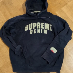 Mörkblå hoodie från Crocker - Mörkblå hoodie från Crocker med texten 'Supreme Denim' på framsidan. Den har en känguruficka och en liten detalj med texten 'Real' på ärmen. Perfekt för en avslappnad stil.