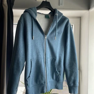 Blå hoodie från Superdry - Säljer en snygg blå hoodie från Superdry i mycket bra skick. Den har en dragkedja framtill och en mysig fodrad insida. Perfekt för höst och vinter! Stor ficka framtill och justerbar huva med snören. Passar både till vardags och chillkvällar. Nypris ca 1200