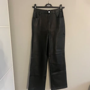 Svarta skinnbyxor från NA-KD - Snygga svarta skinnbyxor från NA-KD med hög midja och raka ben. Byxorna har fickor både fram och bak samt en knapp och dragkedja framtill. Perfekta för en stilren look.