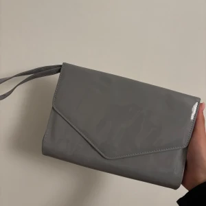 Grå kuvertväska med handledsrem - Snygg och stilren grå kuvertväska med en praktisk handledsrem. Väskan har en enkel design med en tryckknapp för stängning. Perfekt för att bära det viktigaste med stil. Man kan även ta in handledsremmen i väskan. Aldrig använd. 