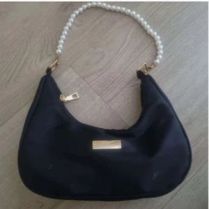 Svart axelväska med pärlhandtag - Snygg svart axelväska med ett unikt pärlhandtag och gulddetaljer. Väskan har en dragkedja för säker förvaring och en elegant design som passar perfekt för olika tillfällen.