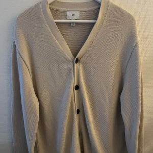Beige kofta från H&M - Säljer en stilren beige kofta från H&M med knappar framtill. Perfekt för lager-på-lager under kyligare dagar. Koftan har en mjuk och bekväm passform med långa ärmar.