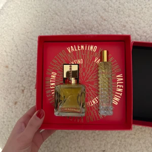 Valentino Voce Viva parfymset - Oanvänd Elegant parfymset från Valentino med doften Voce Viva. Innehåller en fyrkantig flaska med guldfärgad kork och en mindre, mönstrad flaska. Förpackningen är röd med guldtext och designad för att ge en lyxig känsla. Perfekt för den som vill ha en sofistikerad doftupplevelse.