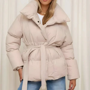 Beige pufferjacka med bälte - Säljer en supermysig beige pufferjacka som är perfekt för kyliga dagar. Den har ett bälte i midjan för en snygg passform och stora, puffiga ärmar. Jackan är långärmad och har en hög krage som skyddar mot vinden. Perfekt för både höst och vinter!