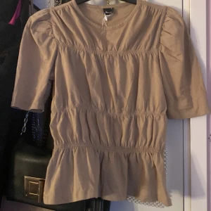 Beige blus med puffärmar - Säljer en söt beige blus med puffärmar och smockdetaljer. Blusen har en volangkant nedtill och trekvartsärmar, vilket ger den en romantisk touch. Perfekt för både vardag och fest!
