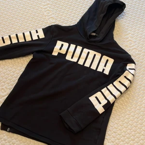 Svart hoodie från Puma - Säljer en svart hoodie från Puma i använt skick. Den har en stor logga på bröstet och ärmarna. DEn är lätt urtvättad i färgen och det finns en liten skada på baksidan, men inget som stör användningen.