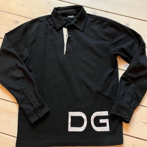Svart pikétröja från D&G - Snygg svart pikétröja från Dolce Gabanna med vita detaljer och stora DG-bokstäver på framsidan. Tröjan har långa ärmar och en krage. Passar som en S-M mer