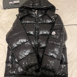 Svart dunjacka från Moncler - Snygg svart dunjacka från Moncler med glansig finish. Jackan har en dragkedja framtill och en praktisk huva. Den är dekorerad med Monclers logotyp på ärmen och har flera fickor med knappar.