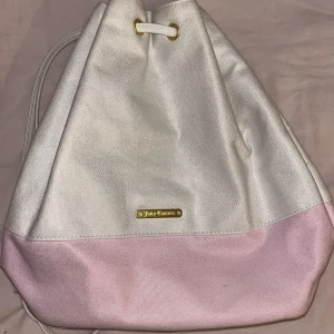 Vit och rosa ryggsäck från Juicy Couture - Ryggsäck i vitt och rosa från Juicy Couture. Väskan har en dragkedja och en rosettdetalj. Insidan är randig i rosa och vitt.