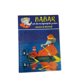Babar och den hemlighetsfulla grottan - En charmig barnbok om elefanten Babar och hans äventyr i en mystisk grotta. Boken har en färgglad framsida med Babar i en båt, klädd i röd kostym och gul krona. Perfekt för unga läsare som älskar spännande berättelser och fantasifulla illustrationer.