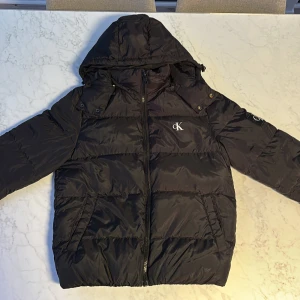 Svart pufferjacka från Calvin Klein Jeans - Snygg svart pufferjacka från Calvin Klein Jeans med huva och dragkedja. Jackan har märkets logga på bröstet och insidan. Perfekt för kyliga dagar med sin vadderade design. Pris kan diskuteras