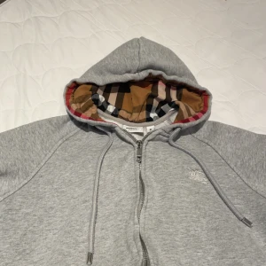 Burberry kofta  - Snygg grå hoodie från Burberry med klassiskt rutigt mönster i huvan. Tröjan har dragkedja framtill och justerbara snören vid huvan. Perfekt för en stilren och bekväm look. Storleken är s. (Köpte för 1199 säljer för 599)