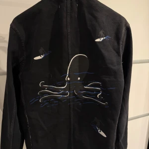 Svart jacka med bläckfiskmotiv från Our Legacy - Säljer en svart jacka från Our Legacy med ett coolt bläckfiskmotiv på ryggen. Jackan har en unik design med ståkrage och knappar framtill. Perfekt för höst och vår, och den är i mycket bra skick. Passar både till vardags och fest!