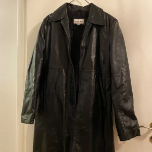 Svart skinnrock från Hollies - Helt ny Snygg svart skinnrock från Hollies i storlek 38. Rocken har en klassisk design med knappar framtill och långa ärmar. Perfekt för en stilren look.
