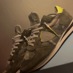  Philippe Model skor  - Säljer ett par snygga sneakers från Philippe Model i camouflage-mönster. Skorna har en cool mix av gröna och beige toner med detaljer i gult. De är i bra skick och har klassisk snörning. Perfekta för en avslappnad stil med en twist!