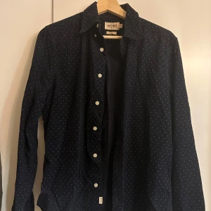 Mörkblå prickig skjorta från East West - Snygg mörkblå skjorta från East West med små vita prickar. Den har en klassisk krage och knappar hela vägen ner. Perfekt för både vardag och fest! Skjortan är i slim fit och har långa ärmar. Passar bra till jeans eller chinos för en stilren look. Storlek S