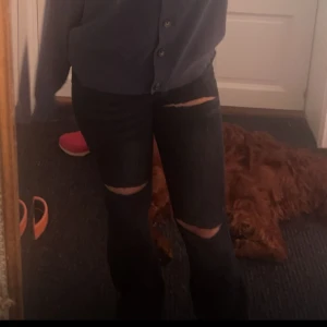 Svarta bootcut jeans Low waist - Säljer ett par svarta jeans med coola slitningar på knäna. Perfekta för en avslappnad och trendig look. De är tighta och passar bra till både vardag och fest. Sköna att ha på sig och lätta att matcha med olika toppar.
