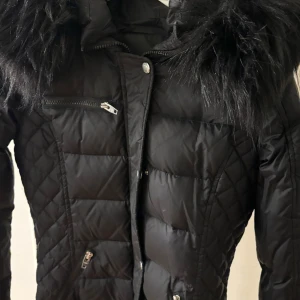 Svart quiltad jacka från Rockandblue - Säljer en snygg svart quiltad jacka från Rockandblue i storlek 38. Jackan har en fluffig fuskpäls på huvan och dragkedjor på fickorna. Perfekt för kyliga dagar och ger en cool look. Passar både till vardags och fest!