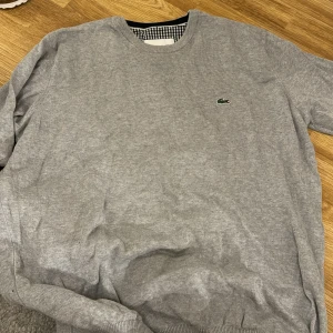Grå tröja från Lacoste - Säljer en stilren grå tröja från Lacoste i mycket bra skick. Den har en klassisk rund hals och det ikoniska krokodilmärket på bröstet. Perfekt för både vardag och lite finare tillfällen. Passar bra till höst och vår!