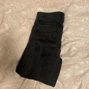 Svarta jeans från Nudie Jeans - Säljer ett par svarta jeans från Nudie Jeans i storlek 31/34. De har en klassisk femficksdesign med knappar i gylfen. Jeansen är i bra skick och passar perfekt för både vardag och fest. De är tillverkade i slitstarkt denim och har en snygg, mörk tvätt. Modell Grim Tim, nypris 1600kr.