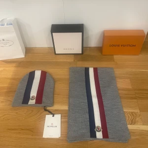 mössa och halsduk från Moncler - Helt nytt och oanvänt Snyggt set med grå mössa och halsduk från Moncler. Båda har en stilren design med ränder i blått, vitt och rött. Perfekt för att hålla sig varm och trendig under de kallare månaderna. Mössan har en broderad Moncler-logga framtill. Passar perfekt för både vardag och mer uppklädda tillfällen.
