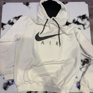 Nike hoodie - Säljer en vit hoodie från Nike med svart logga och texten 'AIR' på framsidan. Den har en justerbar huva med svarta snören och ribbade muddar vid ärmslut och nederkant. Perfekt för en avslappnad stil hemma eller träning. 