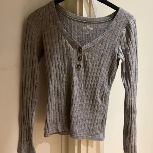 Grå ribbad tröja från Hollister - Säljer en snygg grå ribbad tröja från Hollister i storlek S. Den har en v-ringad design med tre knappar framtill och långa ärmar. Perfekt för både höst och vår, och den är supermjuk och bekväm att ha på sig. Passar perfekt till jeans eller kjol för en avslappnad look.