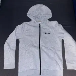 Säljer en stilren grå hoodie från BOSS i storlek S. Den har en dragkedja framtill och en bekväm huva. Perfekt för både höst och vår, med långa ärmar och mjukt material. Passar till både vardag och avslappnade tillfällen.